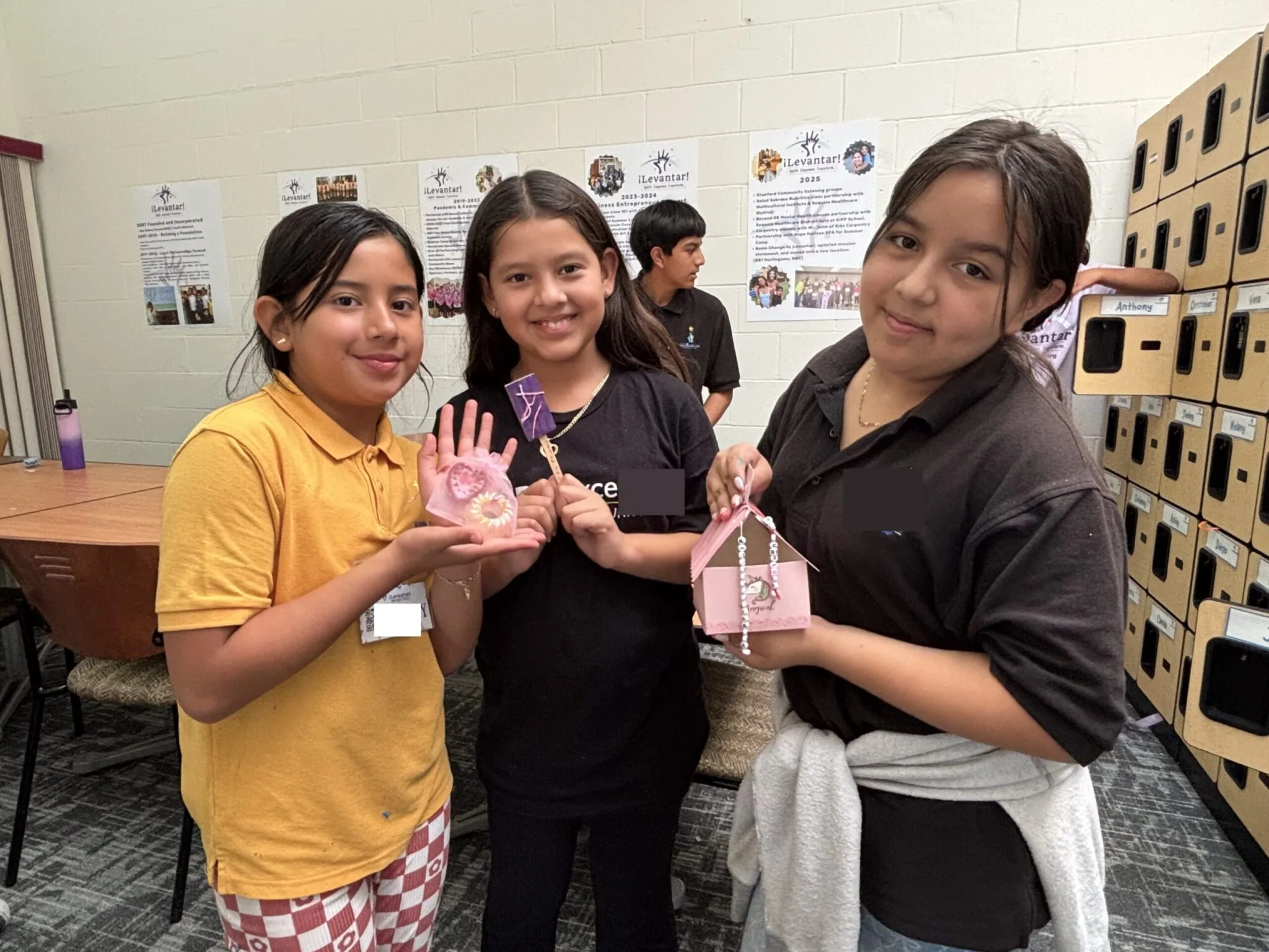 Three upper elementary aged girls of hispanic descent display a handmade craft in their classroom. They have brown hair and are wearing school uniforms with the names covered for privacy.